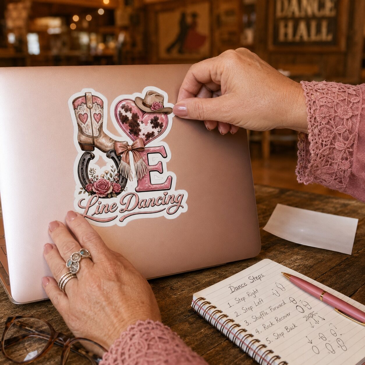 Woman applying a pink LOVE Line Dancing kiss-cut sticker to a rose-gold laptop at a country dance hall