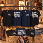 Black and navy Pour Me a Drink Let’s Dance cotton T-shirts hanging in a rustic stable with front and back designs, cowboy boots, hat and horse